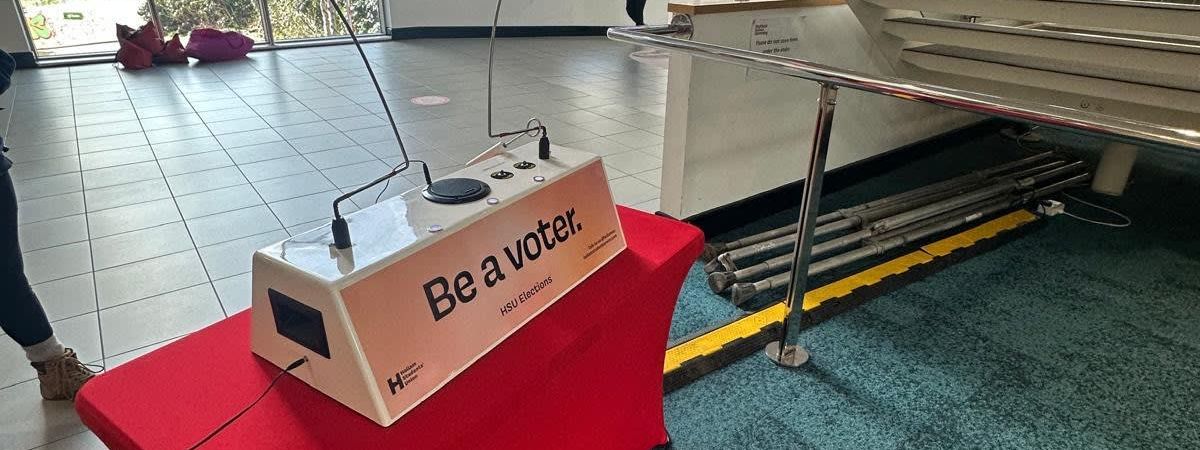 Beat the Buzzer steady-hand game on a red-skirted table branded "Be a voter — HSU Elections" in a university foyer