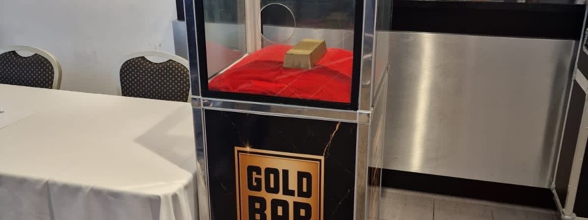 Gold Bar Challenge plinth with a clear-fronted prize chamber containing a gold bar on a red cushion, set up beside a bar