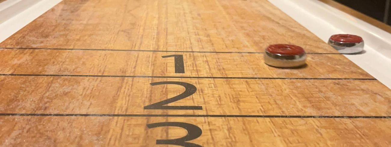 Two men playing shuffleboard in a warm indoor setting, viewed from low angle down the length of the table