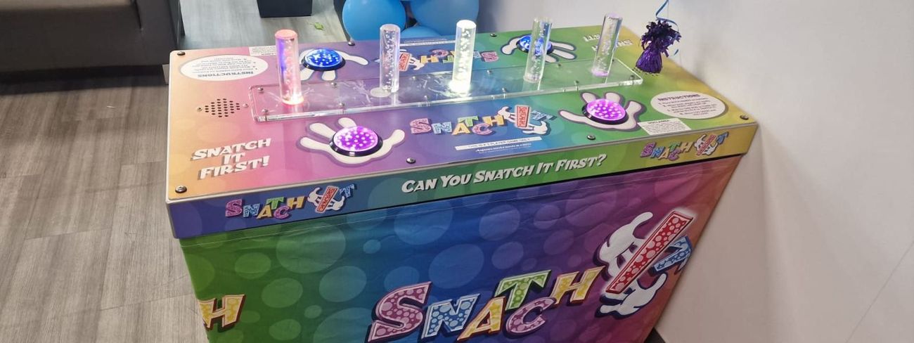 Snatch It game with rainbow branding lit up at an event next to a balloon column