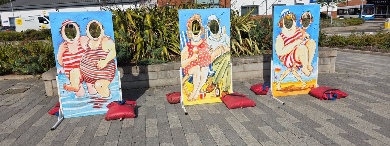 Three beach-themed face-in-the-hole photo cut-out boards standing on metal stands with red sandbag weights on a paved outdoor area