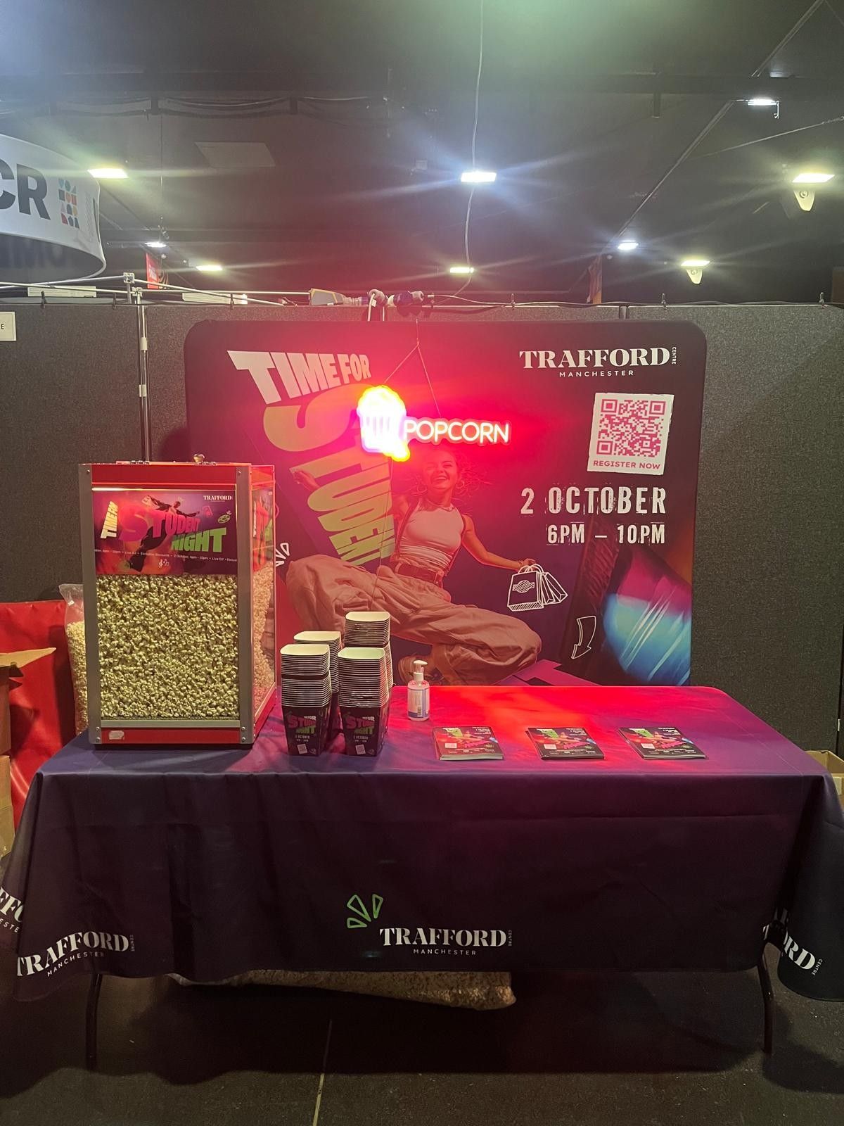 Glass-cabinet popcorn machine on a branded stand with a hanging neon popcorn sign, printed tubs and a QR code backdrop