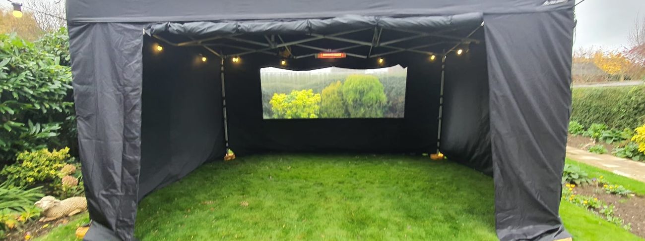 Black 3x4.5m gazebo set up on a garden lawn with festoon lights, a heater and a clear window panel