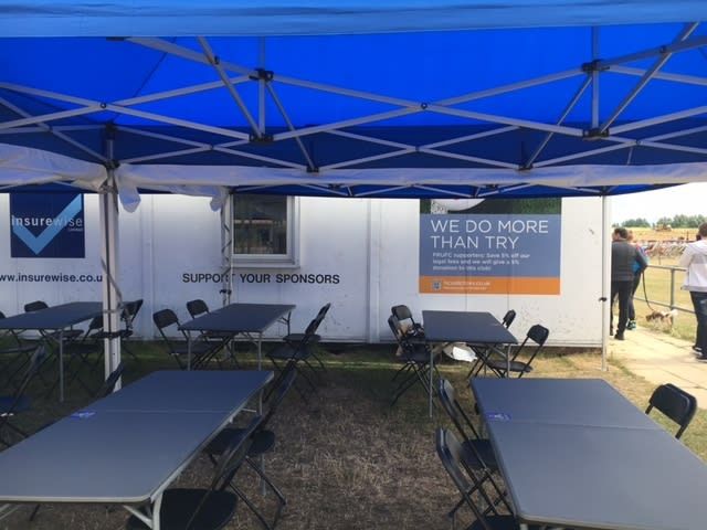 6ft foldable tables and chairs set up under a sponsor-branded blue gazebo at a sports ground