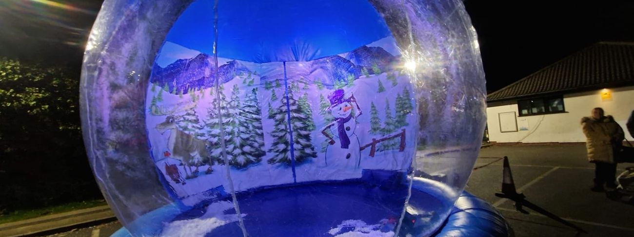 Walk-in giant inflatable snow globe lit with blue interior lighting at night, painted winter backdrop with snowman and reindeer visible through the clear dome