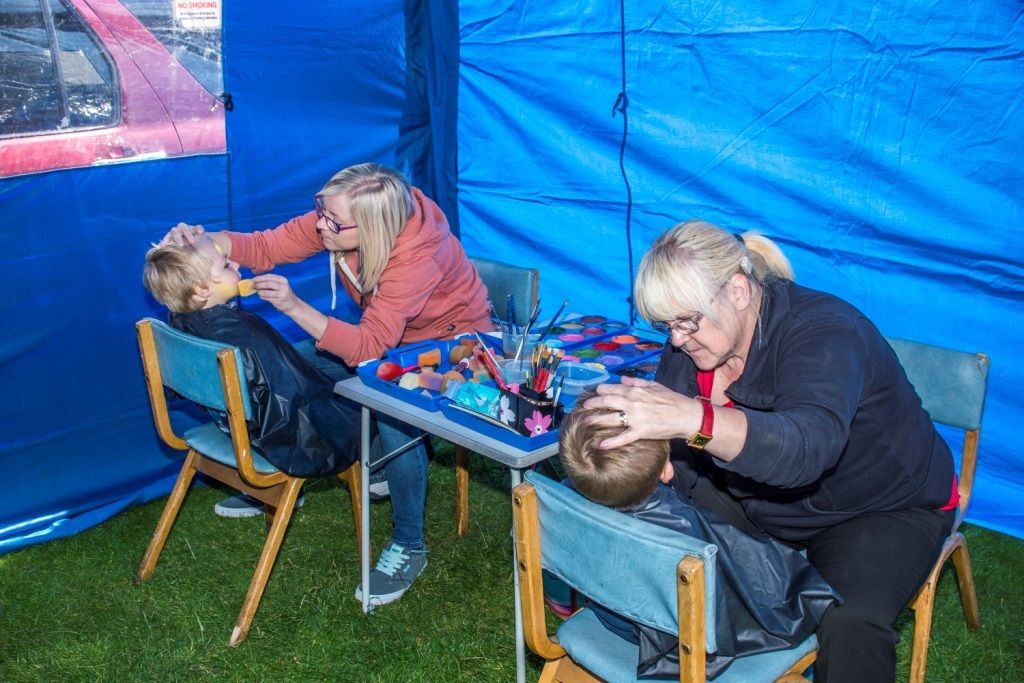 Two face painters working on two children side by side inside a blue pop-up marquee