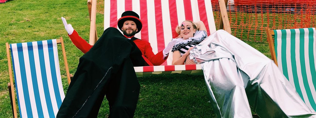 Two performers in costume on a giant red-and-white striped deck chair at an outdoor event, with a DB Entertainment helter-skelter and standard deck chairs in the background