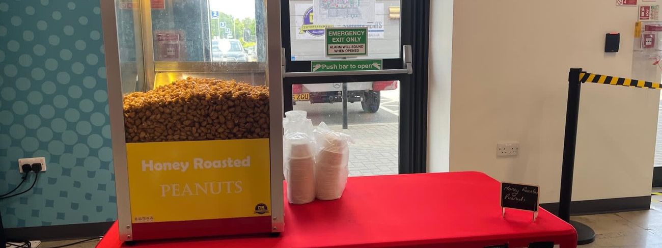 Glass-fronted heated cart with a yellow Honey Roasted Peanuts panel, full of warm peanuts, on a red table with paper portion cups stacked beside it