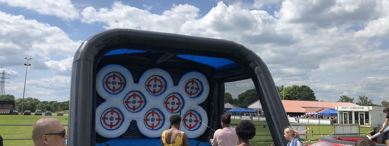 Outdoor community event with inflatable archery arena on grassy field, crowd gathered around watching players