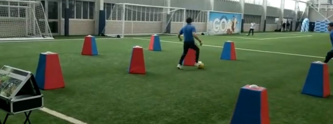 Person in blue polo dribbling a yellow football through red and blue obstacle blocks on an indoor 3G pitch