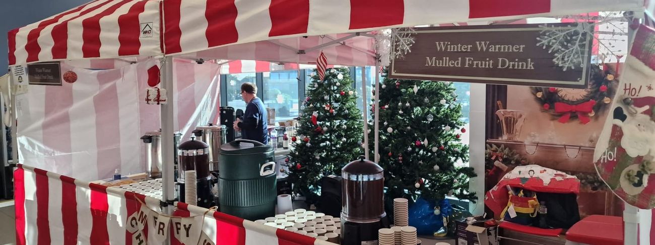 Red and white striped mulled wine stall with "Winter Warmer Mulled Fruit Drink" sign, stainless steel urns, beverage dispensers, stacks of cups and decorated Christmas trees