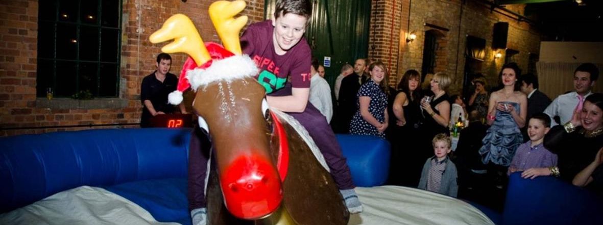 Boy riding the bucking reindeer at a Christmas party in a brick warehouse venue, crowd watching, digital timer visible in the background