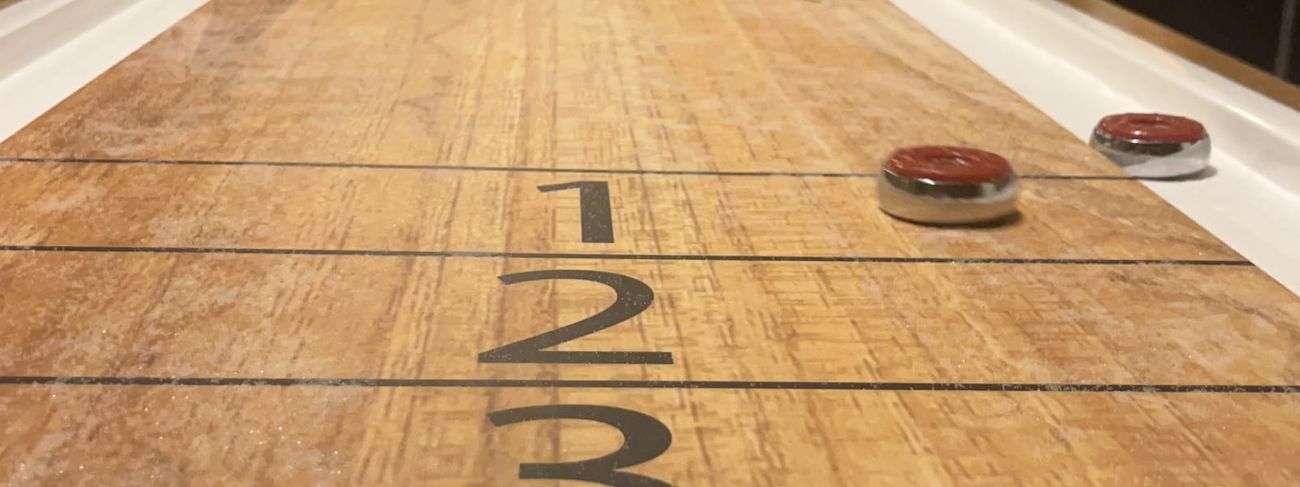 Two men playing shuffleboard in a warm indoor setting, viewed from low angle down the length of the table