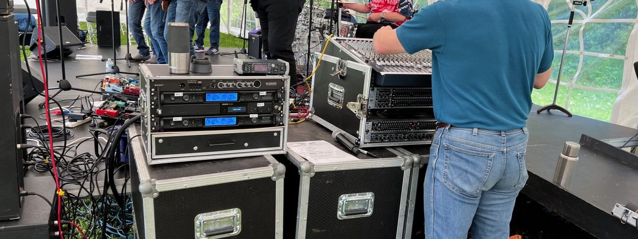 Sound engineer in a teal polo shirt operating a flight-case mixing desk at the side of a marquee stage, with a five-piece band performing behind him