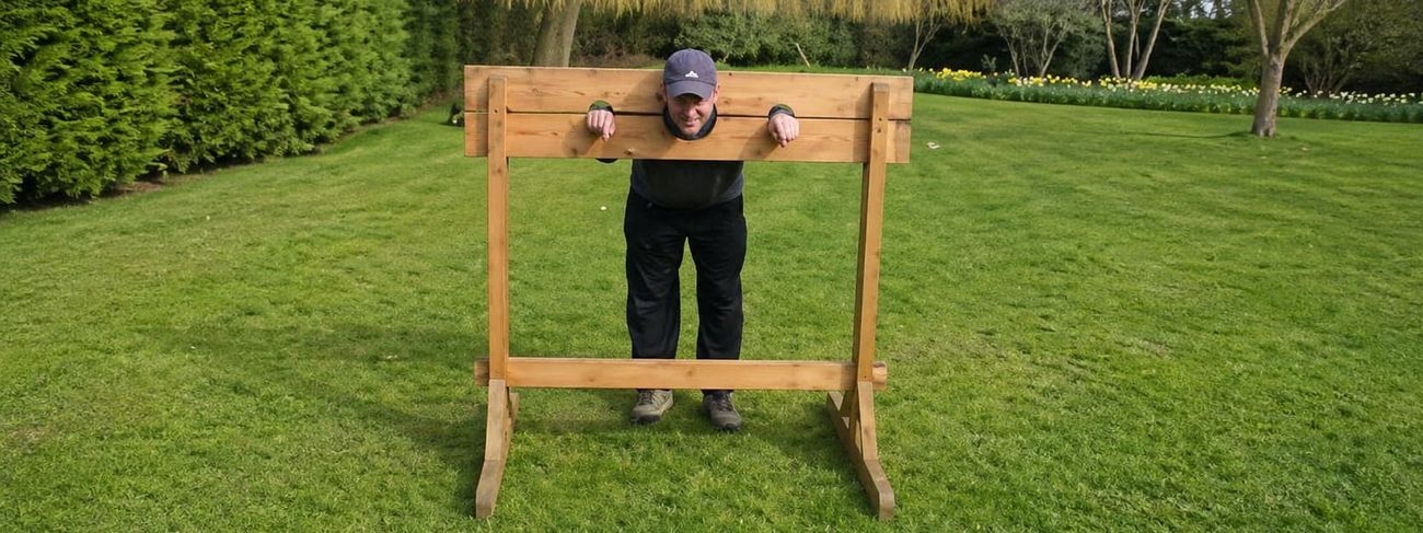 Wooden stocks set up on a lawn in front of a manicured garden with willow trees