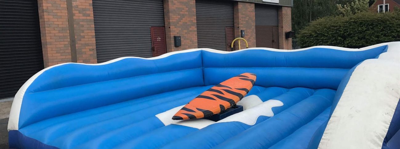 Orange and black tiger-stripe surfboard mounted on a mechanical base in a bright blue inflatable wave surround, photographed outside our warehouse in Rotherham