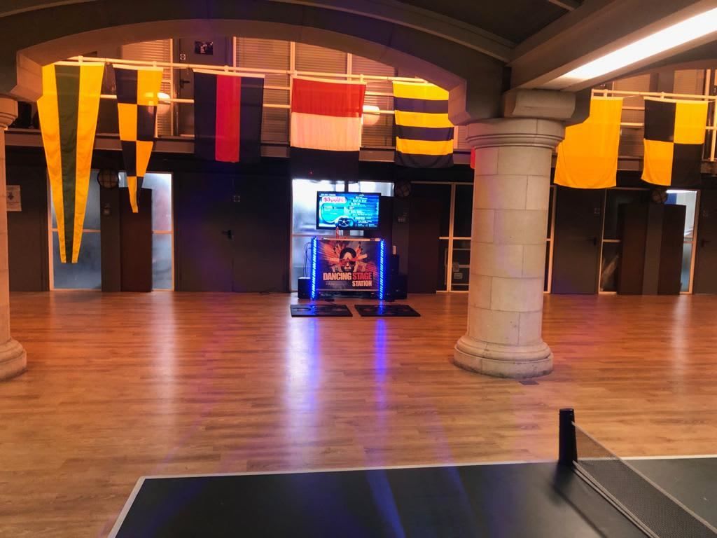 Table tennis table edge in the foreground of a hall set up with a Dancing Stage arcade station and hanging banners