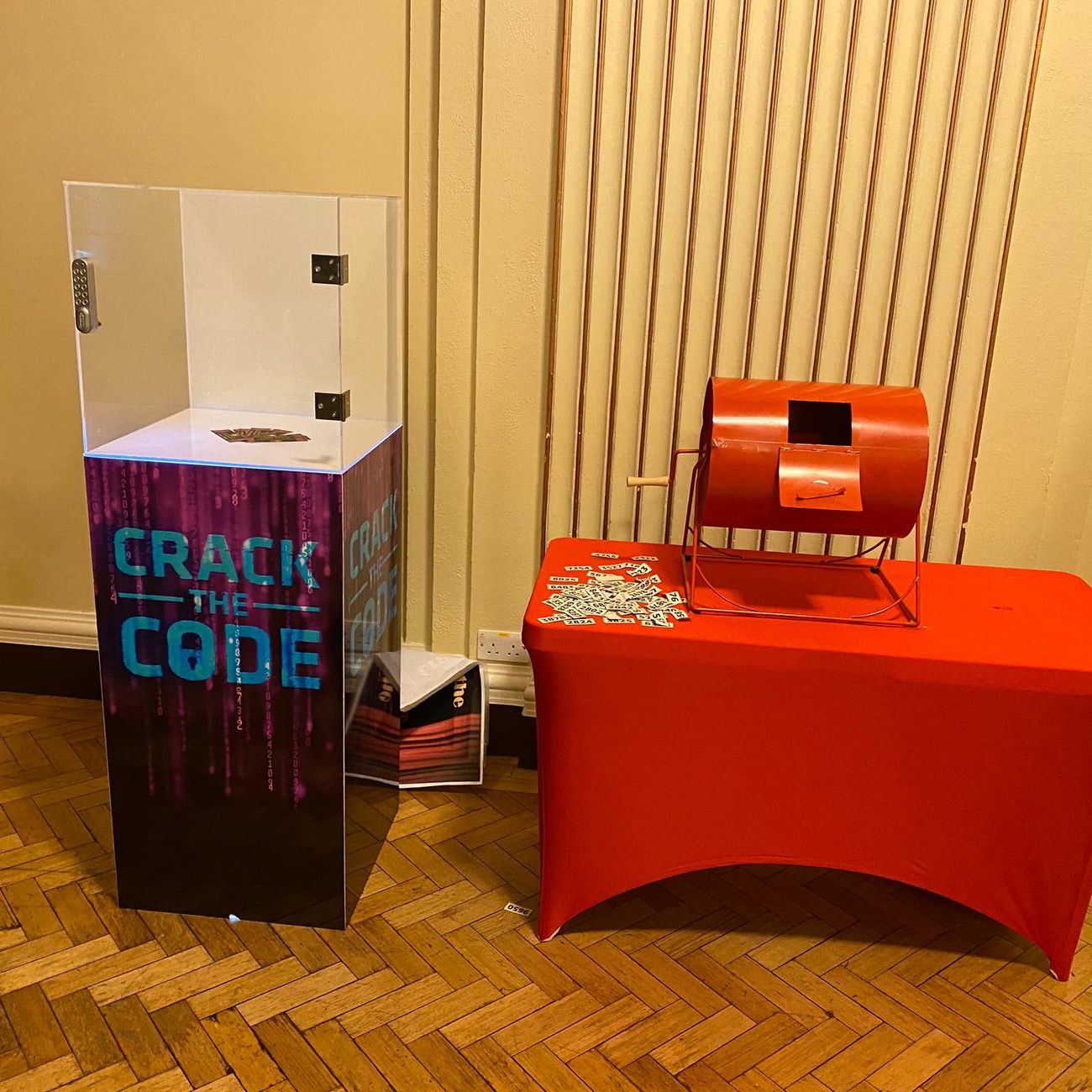 Red tombola drum on a red-skirted table beside a Crack the Code prize plinth at an indoor event